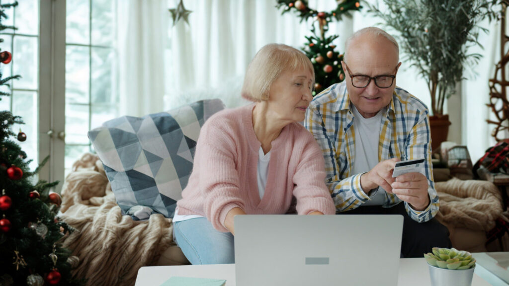 A man and a woman sit on a sofa in front of Christmas decoration, they are looking at a laptop screen and reading the numbers off the back of a credit card